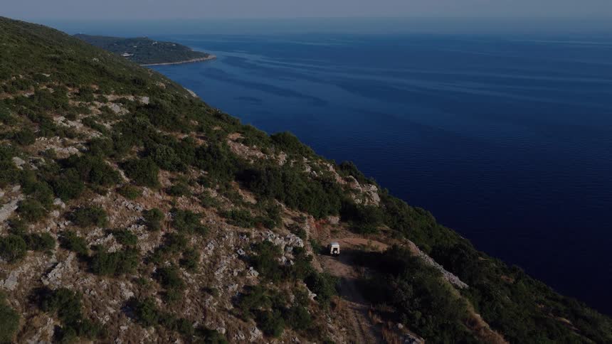An aerial view rises over a rugged, green coastline to reveal a lone camper van parked with a scenic view of the vast blue ocean