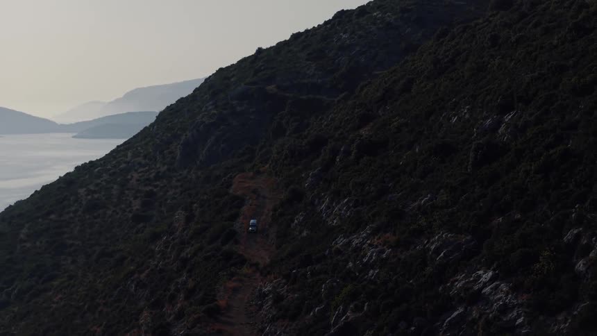 An aerial view shows a white van driving on a winding dirt road along a steep, rugged mountainside overlooking the hazy sea