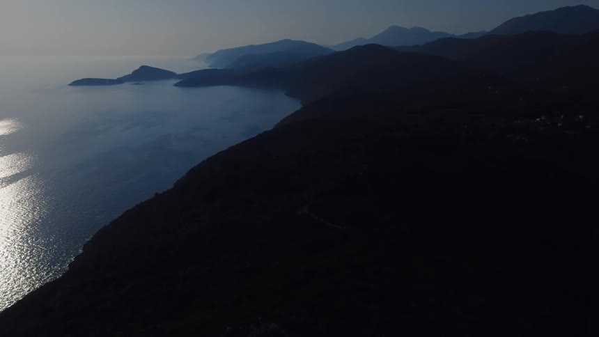 An aerial shot slowly moves over a dark, silhouetted coastline with shimmering light on the calm sea at dawn