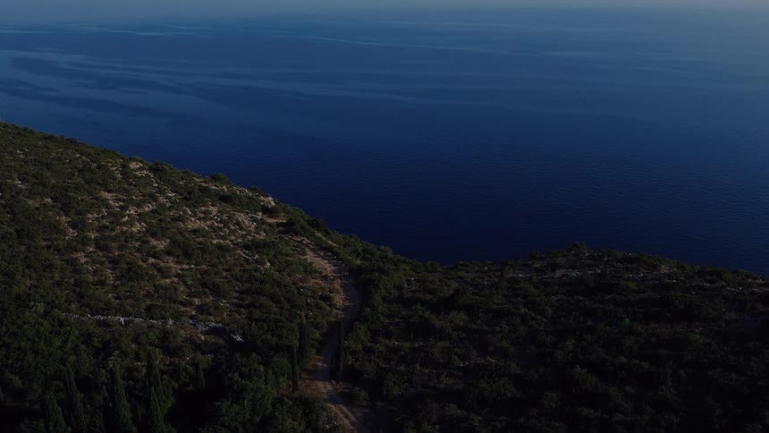 A serene aerial view follows a winding coastal road through a dense green forest overlooking the vast blue sea at sunrise