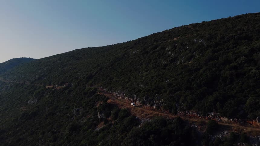 An aerial shot shows a lone white vehicle driving along a remote dirt road on the side of a vast, forested mountain