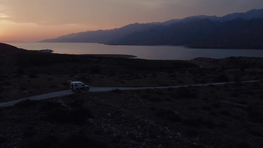 An aerial drone follows an overland vehicle driving on a remote coastal road as the sun sets over the mountains and sea