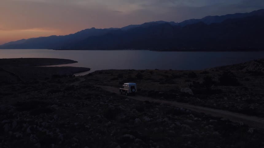 Aerial view of a vintage camper van driving on a remote dirt road along the coast at twilight