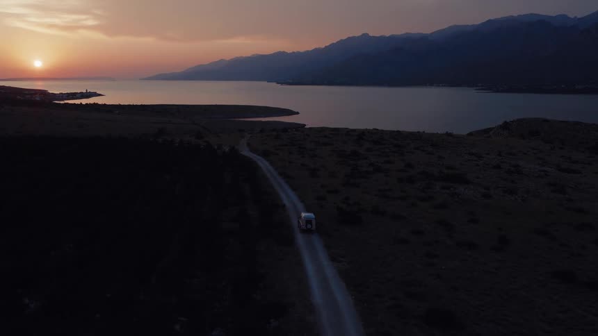 An aerial view of a white camper van driving along a remote coastal dirt road towards the mountains during a beautiful sunset