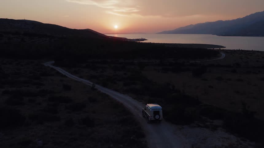 An expedition vehicle drives down a winding dirt road along a rugged coastline as the sun sets over the distant hills