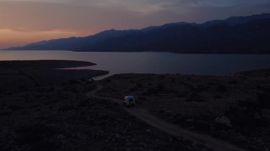 An off-road vehicle navigates a winding dirt road along a rugged, remote coastline as the sun sets behind distant mountains