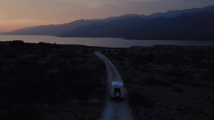 A camper van drives down a winding dirt road toward a tranquil sea and distant mountains during a colorful sunset