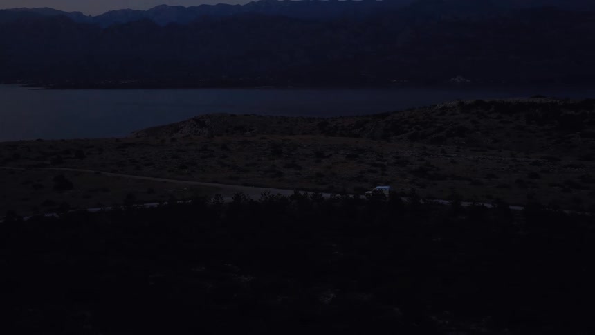 An aerial view of an off-road vehicle driving on a winding road through a mountainous landscape beside a large body of water at twilight
