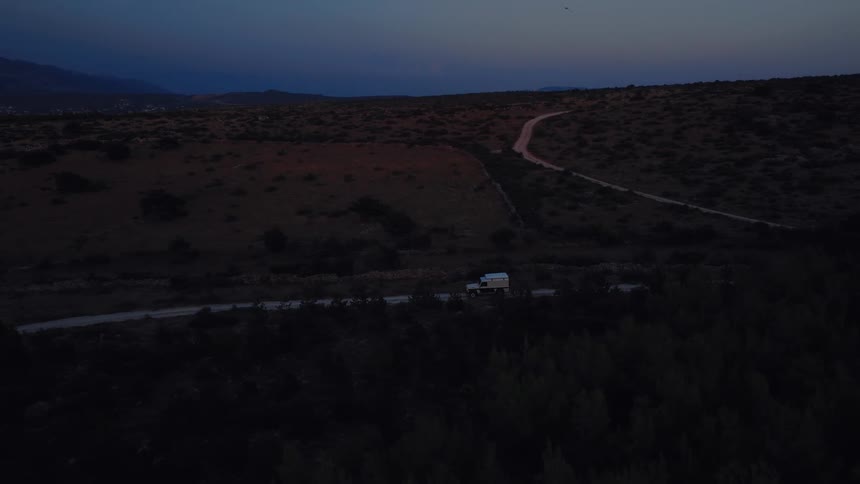 An aerial view captures an off-road vehicle driving along a desolate road through a dark, hilly landscape at twilight