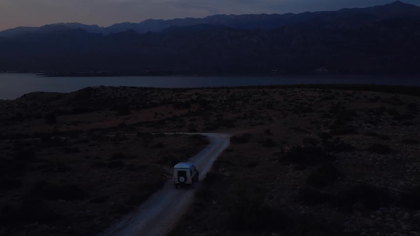 Aerial footage follows a 4x4 camper van driving along a remote coastal dirt road at twilight with mountains in the distance