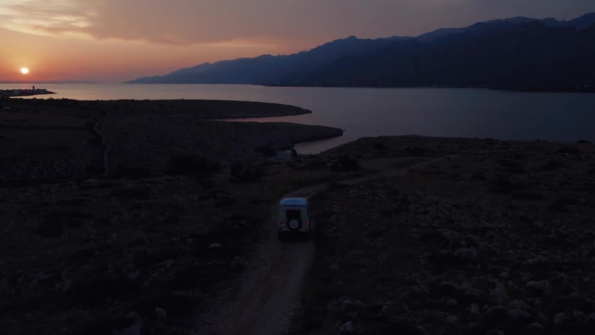 An expedition vehicle drives down a dirt road on a rocky coastline as the sun sets over the mountains and sea