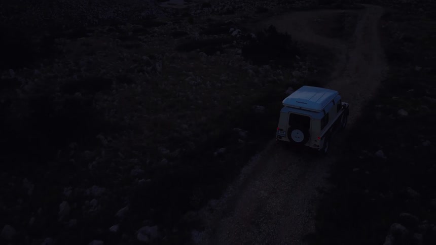 Aerial view follows an off-road camper van driving along a remote, rocky coastal path as the sun sets over the ocean