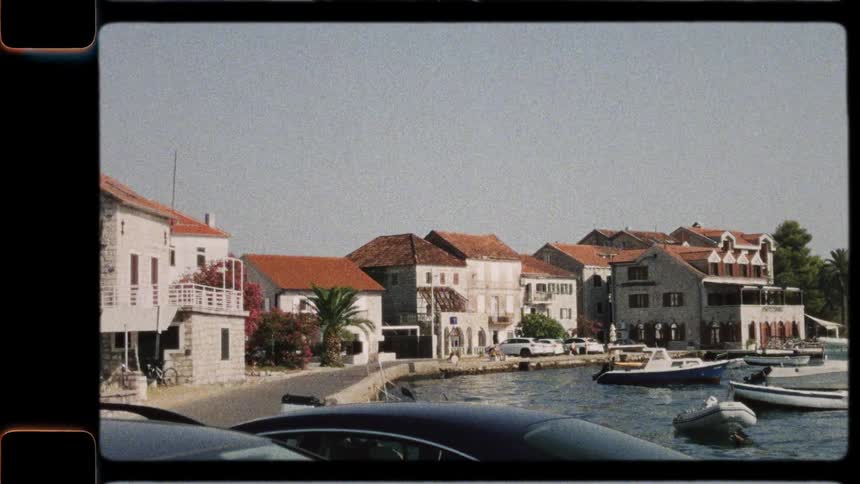 A vintage film recording captures a scenic coastal European town with boats docked in the harbor on a sunny day