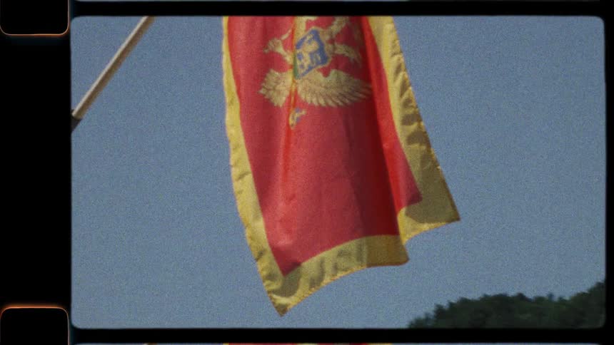 A vintage Super 8mm film shot of the Montenegrin flag waving with a lush mountain in the background