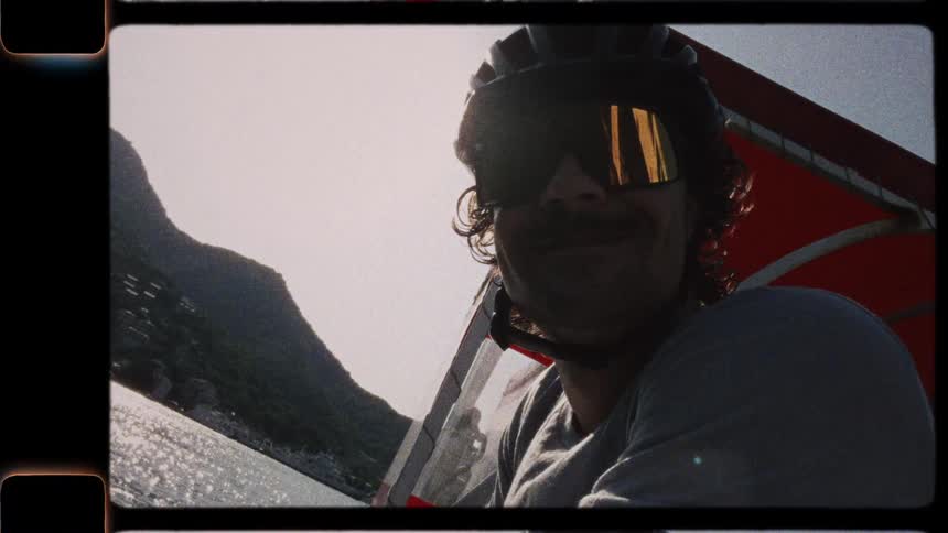 Vintage 8mm film style footage shows a smiling man in sunglasses and a helmet hang gliding over the ocean