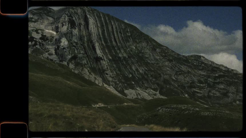 A vintage, archival shot of a large, striated mountain with a narrow road winding at its base