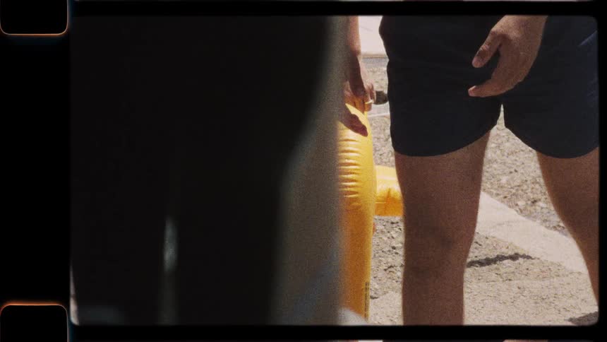 A man in swim trunks picks up a yellow inflatable toy in a nostalgic, vintage-style shot