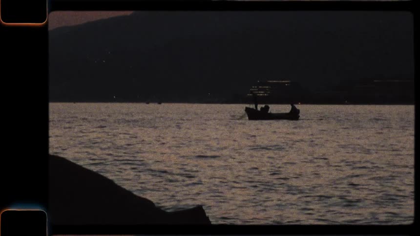 A vintage 8mm film shows the silhouette of a small fishing boat floating on the calm sea at dusk