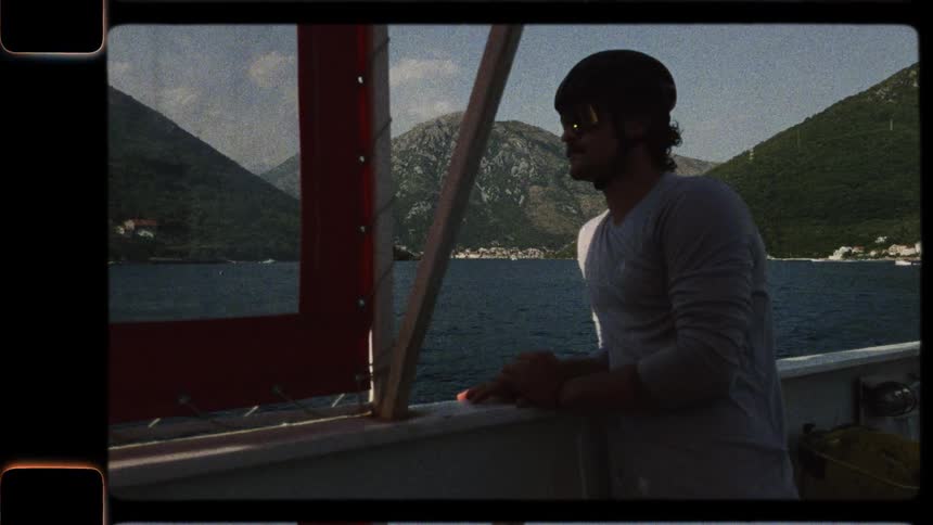 A man wearing a helmet and sunglasses stands on a boat, looking out at the scenic mountains and water in this vintage-style footage