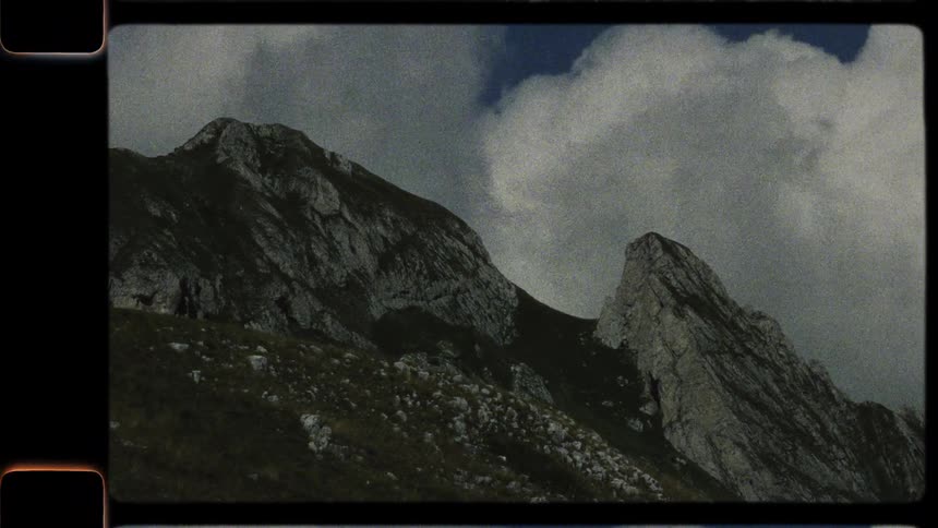 A slow pan across a majestic and rocky mountain range under a cloudy sky, captured on grainy vintage film