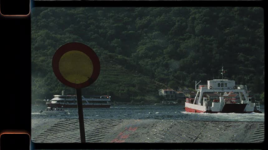 A vintage 16mm film shows a car ferry sailing away from the dock along a scenic, tree-covered coastline