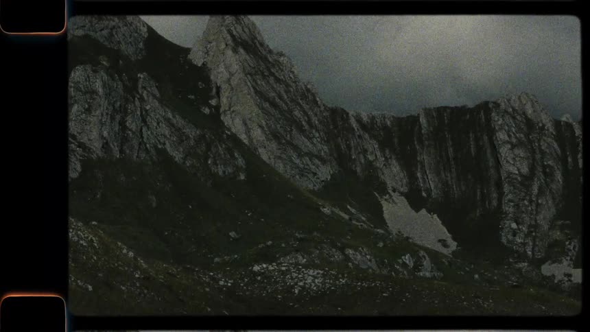 A vintage film-style panning shot reveals the dramatic and rugged peaks of a mountain range under a cloudy sky