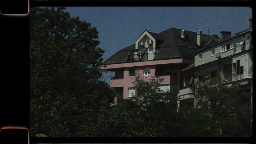 A vintage 8mm film-style shot of a pink apartment building partially obscured by green trees swaying in the wind