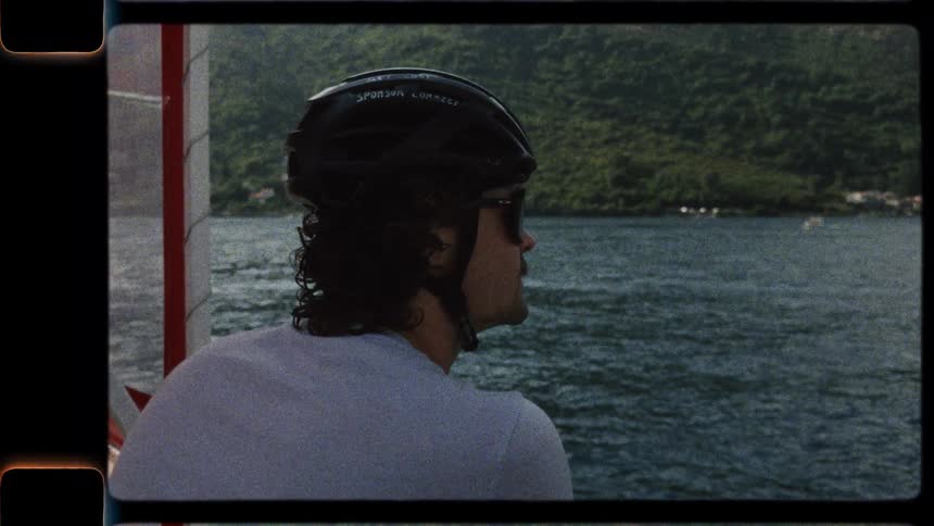 A male cyclist with a helmet and sunglasses enjoys the scenic view from a ferry boat, captured in a vintage film style