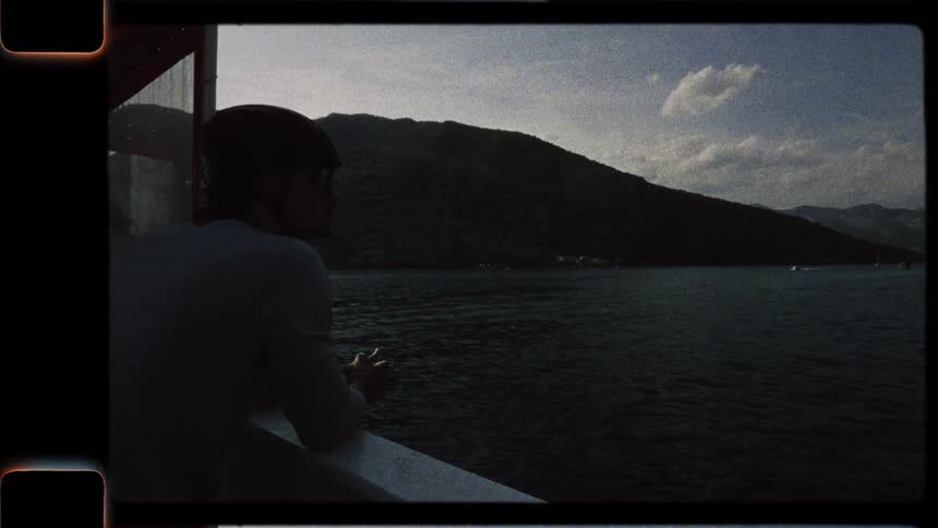 Vintage 8mm-style footage of a man in a helmet looking out at a mountain lake from the deck of a boat