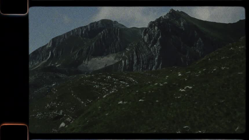 A vintage, 8mm film-style shot captures the majestic and rugged mountain peaks towering over green, rolling hills