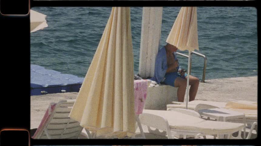 A vintage film captures a man relaxing on a sunny seaside pier next to lounge chairs and umbrellas