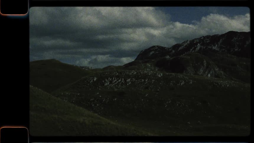 An archival style panning shot reveals a vast, rolling green mountain range under a cloudy sky