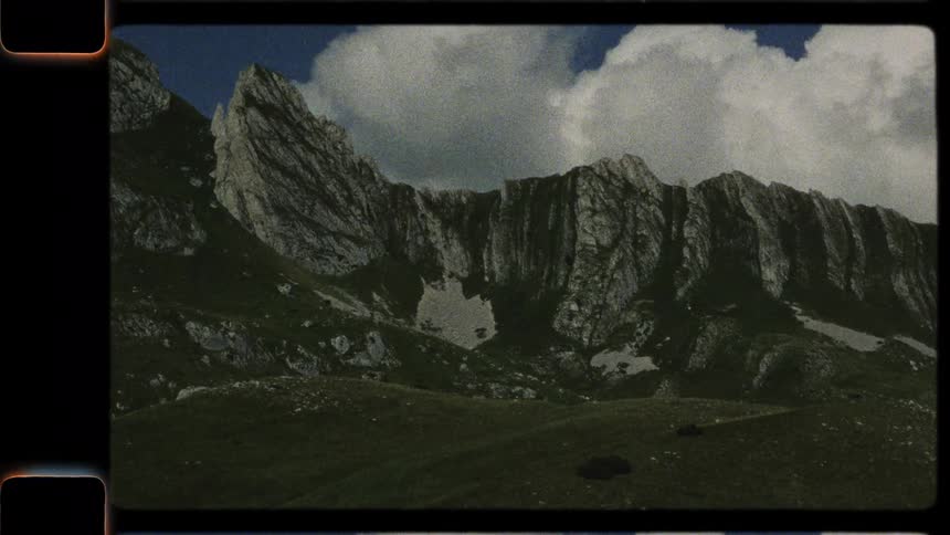 A vintage 8mm film captures a slow, cinematic pan across a majestic and rugged mountain range on a summer day