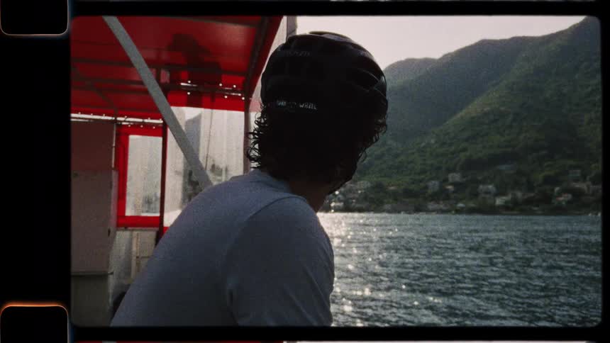 A cyclist wearing a helmet enjoys the scenic mountain and water views from a boat in this vintage film-style footage