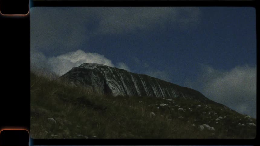A vintage-style shot slowly reveals a grassy hillside below a majestic, striated mountain peak against a partly cloudy blue sky