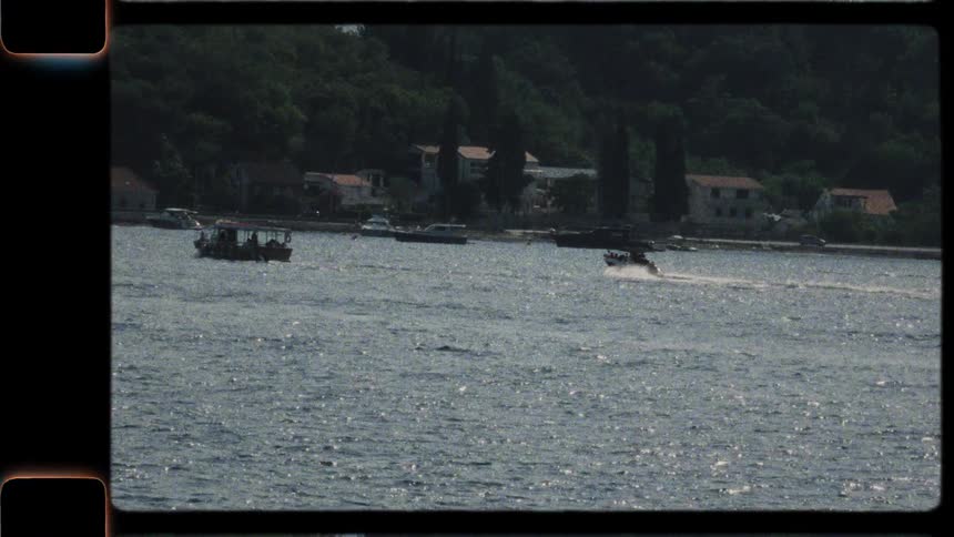 Vintage 8mm-style footage of two motorboats carrying tourists as they speed across a sparkling sea along a lush, green coastline