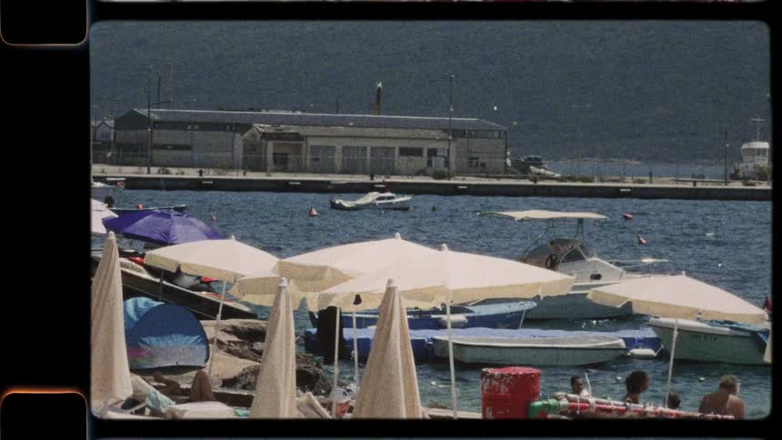 Vintage-style footage pans across a sunny beach with umbrellas and lounge chairs next to a harbor full of boats
