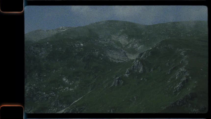 A vintage 8mm film captures a slow pan across a majestic green mountain range with clouds rolling over the peaks and a cabin nestled in the foreground