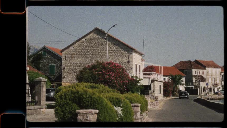 Vintage 8mm film footage pans across a scenic Croatian seaside town, following a car along the harbor and revealing a stone clock tower