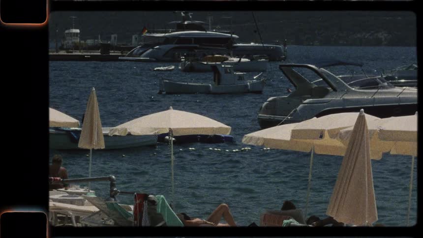Vintage 8mm film captures a sunny day at a busy seaside resort, with people relaxing on lounge chairs and boats anchored in the bay