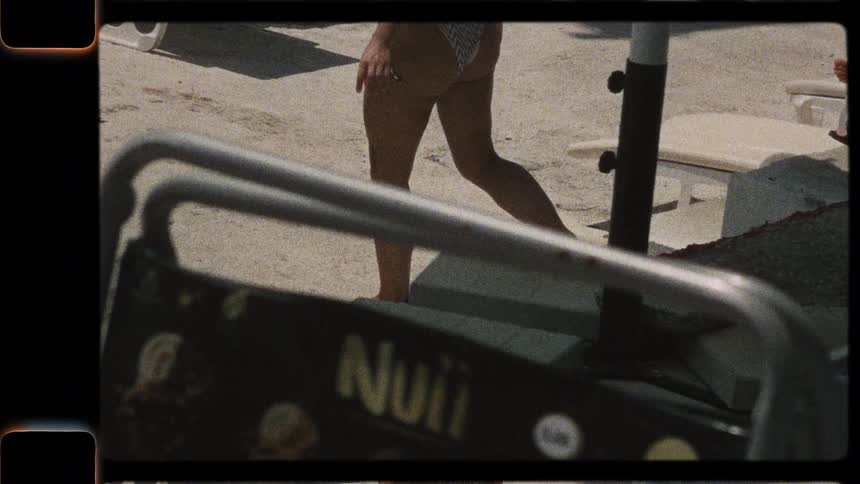 Vintage 8mm style home movie footage of a woman in a bikini walking by a man relaxing under a beach umbrella