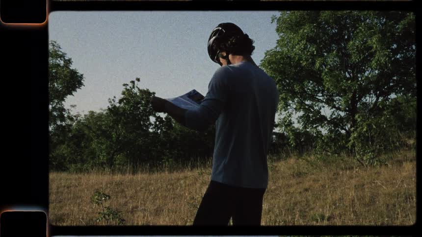 Vintage-style footage shows a male cyclist in a helmet stopping in a sunlit field to unfold and check his paper map