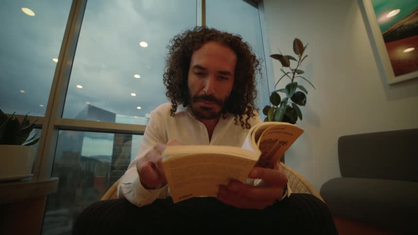 A man with curly hair reads a book in a modern apartment overlooking a cloudy city skyline as the camera slowly pushes in
