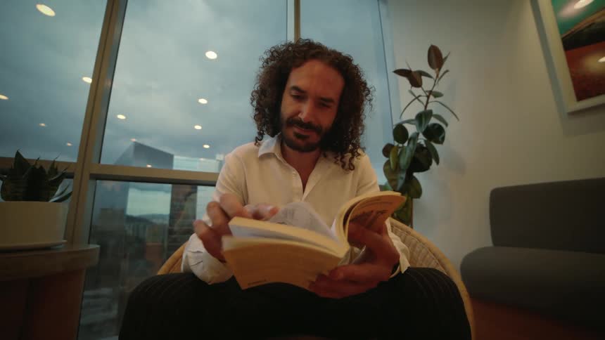 A man with curly hair reads a book while sitting in a modern apartment with a city view at dusk