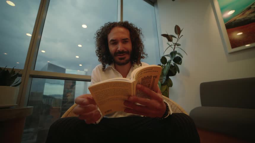 A stylish man with curly hair sits in a modern high-rise and reads a book in slow motion with the city skyline in the background