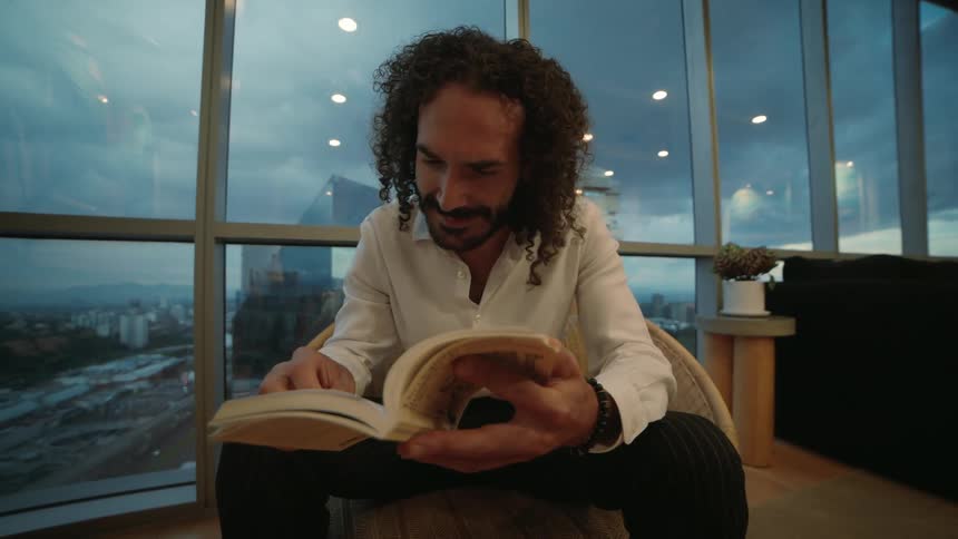 A well-dressed man with curly hair enjoys reading a book in slow motion in a modern high-rise lounge overlooking the city at dusk