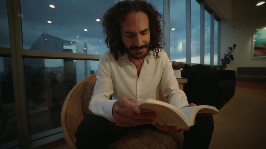 Stylish man with curly hair and a beard reads a book in a wicker chair in a modern lounge overlooking the city at dusk