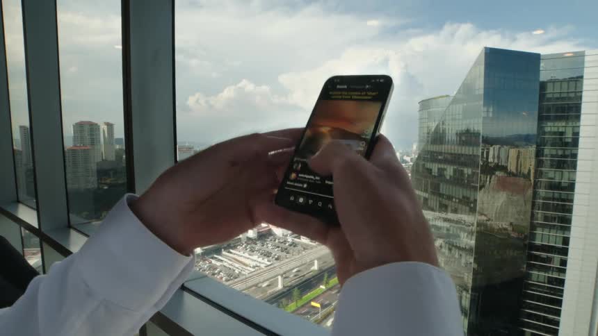 An over-the-shoulder view of a person scrolling through a social media feed on their smartphone in a high-rise office overlooking the city