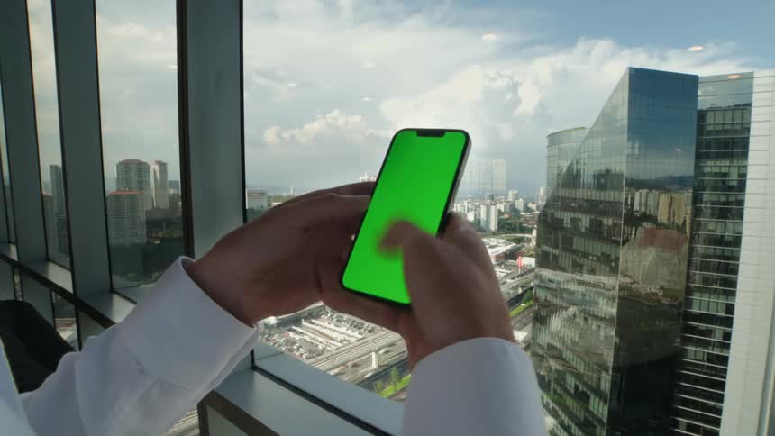 A businessman scrolls on a green screen smartphone while standing by a window in a high-rise office with a cityscape view