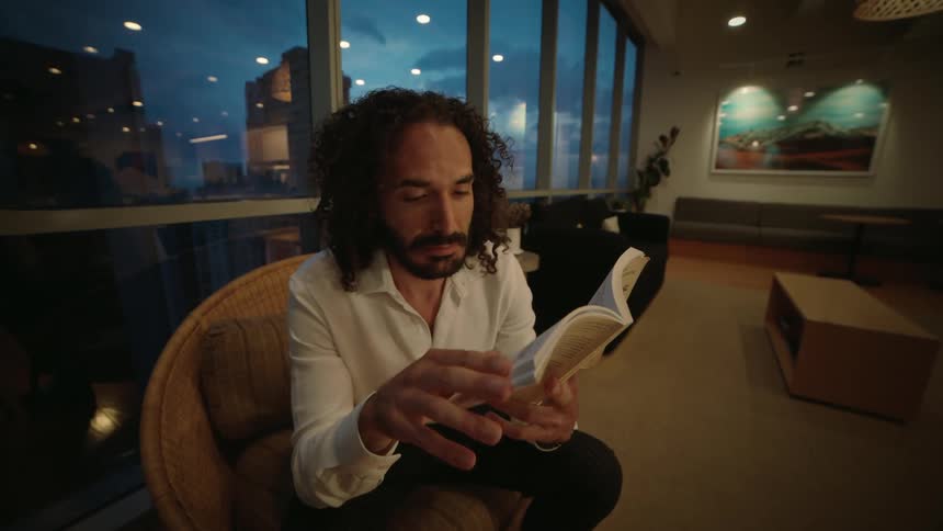 A man with curly hair reads a book intently while sitting in a cozy chair in a modern lounge at dusk